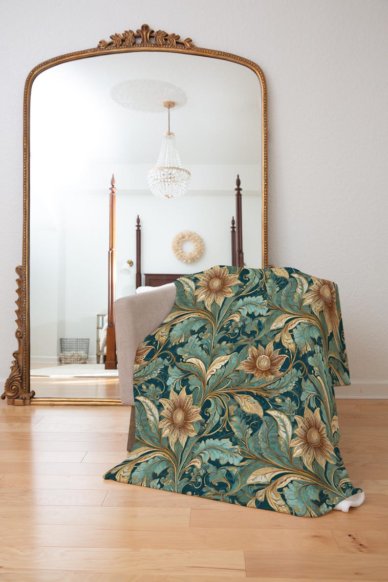 May include: A teal blue throw blanket with a gold and brown floral pattern featuring sunflowers. The blanket is folded and lying on a wooden floor in front of a large ornate gold mirror.