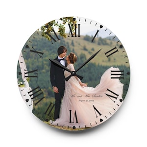 May include: A round, white clock with Roman numerals and black hands. The clock face features a photo of a couple in wedding attire, standing in a field with a mountain in the background. The text "Mr. and Mrs. Sanders August 31, 2023" is printed below the couple.