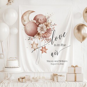 May include: A white backdrop with the text "Love is in the air" in black cursive script.  The backdrop is decorated with a cluster of blush pink balloons, a crescent moon, and a floral arrangement.  The text "Stacey and William August 13, 2022" is printed below the main text.