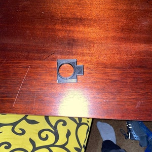 May include: A black plastic 3D printed object with a circular opening in the center. The object is on a wooden surface.