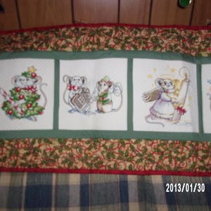 May include: A Christmas-themed fabric panel featuring embroidered mice in festive scenes. The mice are depicted with holiday decorations, including a Christmas tree, angel wings, and holding a candle. The panel has a green border with a holly and berry pattern.