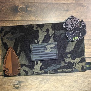 May include: A black and green camouflage pouch with an American flag patch and a brown arrowhead patch. The pouch has a black and white patch with the text "ZFD" and the word "MULTICAM®" printed on the front. A black tag with the text "DownRange" is attached.