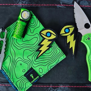 Op de afbeelding: Een verzameling EDC (Everyday Carry) items, waaronder een groen vouwmes, een kleine zaklamp, een multitool, een groene zakdoek met een topografisch patroon en twee oogvormige patches met bliksemaccenten. Op een zwarte ondergrond.