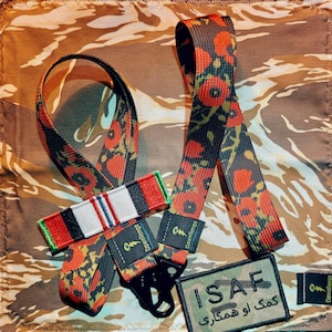 May include: A set of black and red poppy-patterned straps with a black clasp and a patch that reads "ISAF" in white letters. The straps are on a camouflage background.