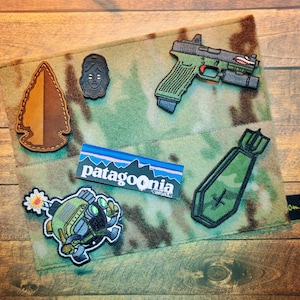 Può includere: Una toppa in tessuto mimetico con varie toppe ricamate. Le toppe includono una punta di freccia marrone, un volto stilizzato, una pistola verde e grigia, una bomba, un robot dei cartoni animati e un logo Patagonia.