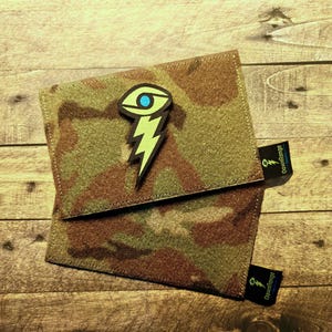 Könnte beinhalten: Quadratische Tasche mit Tarnmuster und einem Blitz- und Augen-Patch. Der Patch ist grün, schwarz und blau. Die Tasche hat zwei schwarze Etiketten mit dem Text "Downrange".