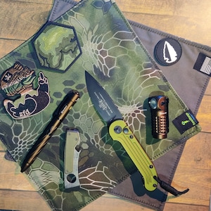 May include: A green and brown camouflage fabric with a black knife, a brown pen, a small multi-tool, and a brown and green patch with a topographic map design.