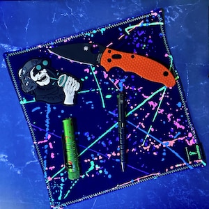 May include: A black and orange folding knife, a black pen, and a green flashlight rest on a dark blue, splatter-painted cloth. A patch of a skull in a helmet holding a spray can is also on the cloth.