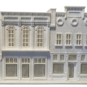 May include: A white 3D printed model of a three-part commercial building facade with brick detailing, arched windows, and storefront displays. The text Building #1 is printed in a box in the upper left corner of the image.