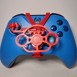 May include: A blue Xbox controller with a red 3D printed steering wheel attachment. The steering wheel has a textured surface and a small red knob in the center.