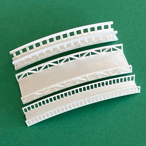 May include: Three white plastic model bridges with different designs. The top bridge has a simple railing, the middle bridge has a lattice design, and the bottom bridge has a more detailed railing.