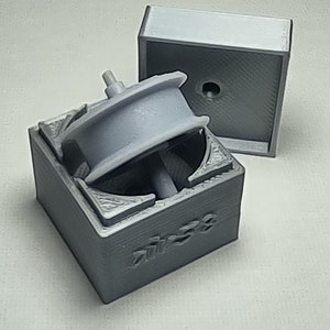 Könnte beinhalten: Eine graue, im 3D-Druck hergestellte Box mit einem sich drehenden Rad im Inneren. Die Box ist geöffnet und das Rad ist sichtbar. Die Box trägt die Aufschrift "DIESEL" an der Seite.