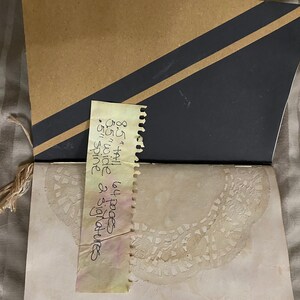 May include: A brown paper notebook with a black and brown striped cover. The notebook is open to a page with a lace design. A yellow note is attached to the cover with the text: "8 1/2 x 11 64 pages 65 wide spine & signatures".