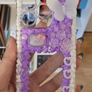 May include: A clear phone case with a purple and white design. The case has a white border with a pearl-like beading. The center of the case is purple with a white butterfly, a white flower, and a white Hello Kitty character.