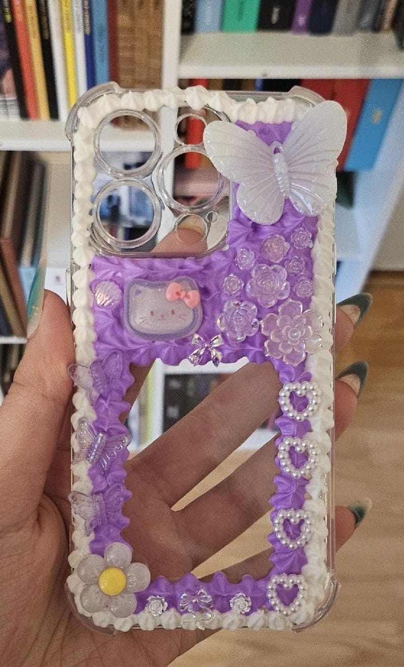 May include: A clear phone case with a purple and white design. The case has a white border with a pearl-like beading. The center of the case is purple with a white butterfly, a white flower, and a white Hello Kitty character.