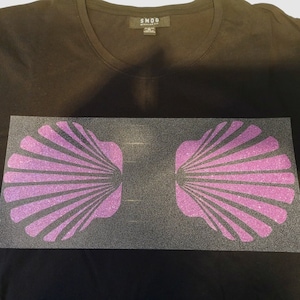 May include: Black crew neck t-shirt with a glittery purple seashell design. The design is centered on the shirt and features two symmetrical seashell shapes. The shirt is made of a soft material and is suitable for casual wear.