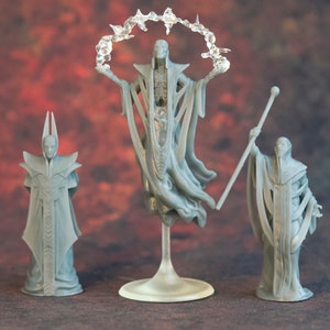 May include: Three gray fantasy figurines. The central figure is a skeletal being with outstretched arms, a halo, and flowing robes. The other two figures are robed characters, one with a staff and the other with a tall, pointed headdress. The figurines are on a gradient background.