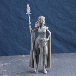 May include: A gray 3D printed figurine of a woman wearing a long dress and a cloak, holding a spear. She has a crown on her head and a necklace around her neck.