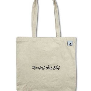 May include: A beige canvas tote bag with black handles and the text "Manifest that Shit" printed on the front.