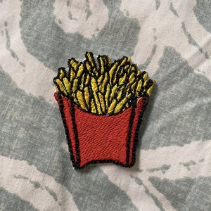 May include: A red and yellow embroidered patch of a cartoon french fry container with fries inside.