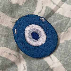 May include: A blue embroidered evil eye charm with a white and blue eye design. The charm is on a white and blue patterned fabric background.