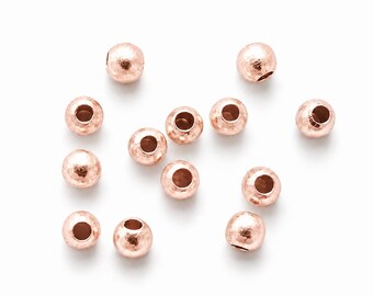 May include: A collection of twelve small, spherical beads with a metallic rose gold finish. Each bead features a central hole for threading and a textured, slightly hammered surface that reflects light, creating a subtle, uneven appearance.