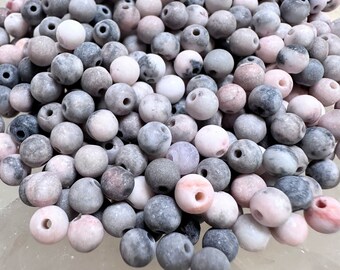 May include: A pile of round, drilled beads in shades of gray, pink, and white. The beads have a matte finish and vary in color intensity, creating a natural, textured appearance. These beads are suitable for jewelry making.