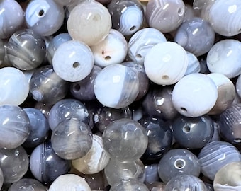 May include: A collection of round, polished beads in shades of gray, white, and tan. The beads have a marbled appearance with visible banding and a smooth, glossy finish. Each bead has a small hole for stringing.