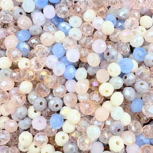 May include: A close-up shot showcases a collection of faceted beads in various pastel colors. The beads are a mix of light pink, pale blue, cream, and iridescent tones. Each bead has a hole for stringing, suggesting their use in jewelry making or crafts.