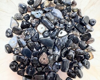 May include: A collection of polished black and gray gemstone beads with drilled holes. The beads vary in size and shape, with some displaying natural patterns and textures. The beads are arranged on a light-colored, textured surface.