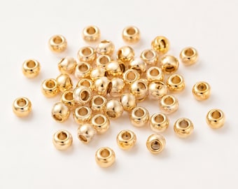 May include: A collection of small, round, metallic gold beads with a smooth, polished finish. Each bead features a central hole for threading, and they are scattered in a loose pile, showcasing their reflective surface and uniform shape.