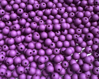 May include: A large collection of small, round, matte purple beads. Each bead features a single, centered hole for threading. The beads are uniform in size and color, creating a dense, textured surface of spherical shapes in a vibrant violet hue.