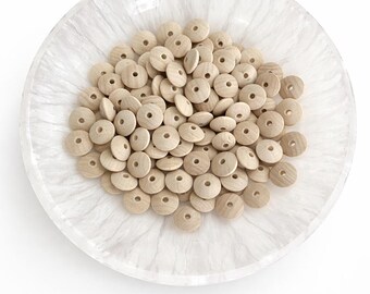 May include: A collection of small, light-colored wooden beads with a hole in the center, displayed in a white, marbled-effect bowl. The beads are round and uniform in size, perfect for crafting or jewelry making.