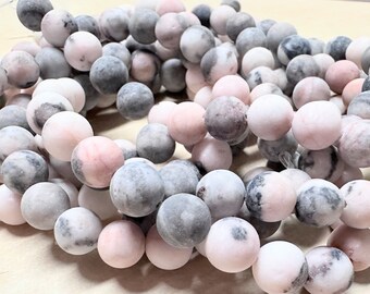 May include: Close-up of a strand of round beads in various shades of pink, gray, and white. The beads have a matte finish and appear to be made of stone or a similar material. The beads are strung together, suggesting they are for jewelry making.