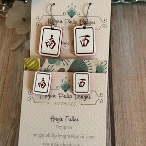 May include: White and red earrings with a Mahjong tile design. The earrings have a rectangular shape with a white background and red border. The earrings are hanging on a white card with text. The card has the designer's name and contact information.