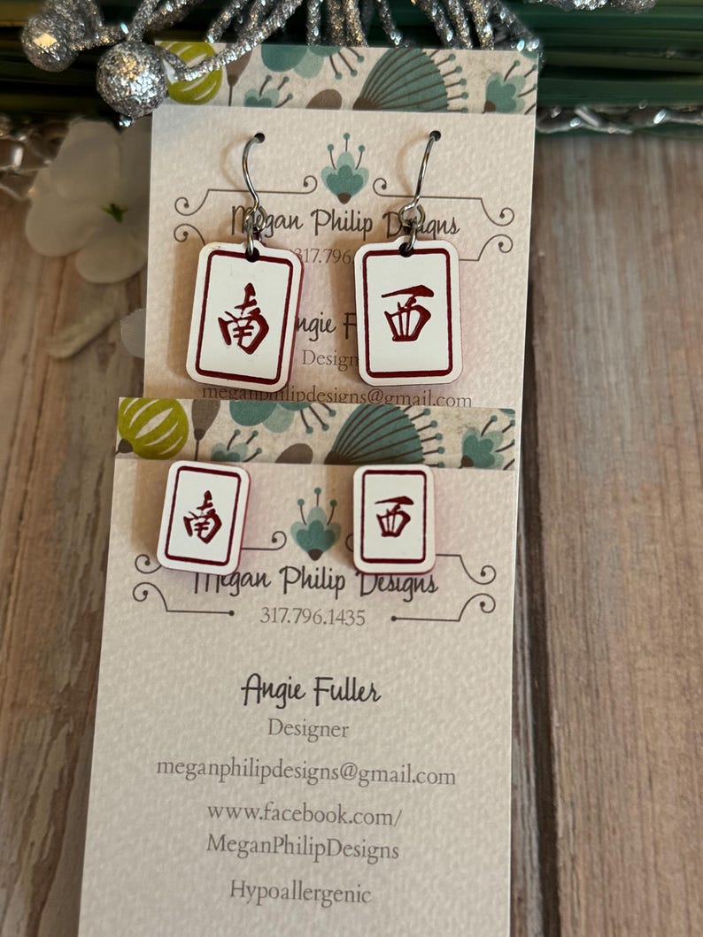 May include: White and red earrings with a Mahjong tile design. The earrings have a rectangular shape with a white background and red border. The earrings are hanging on a white card with text. The card has the designer's name and contact information.