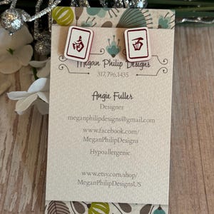 May include: White stud earrings with a red and black Mahjong tile design. The earrings are displayed on a business card with the text "Megan Philip Designs" and contact information. The card is on a wooden surface.