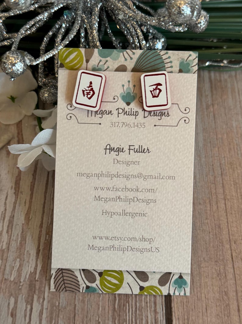 May include: White stud earrings with a red and black Mahjong tile design. The earrings are displayed on a business card with the text "Megan Philip Designs" and contact information. The card is on a wooden surface.