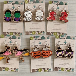 May include: Six pairs of Halloween-themed earrings on display cards. The earrings are made of wood and feature various characters, including Frankenstein, a mummy, a devil, a witch, a flamingo, and a pumpkin mug.