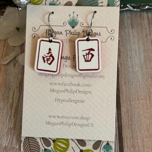 May include: White rectangular earrings with red Chinese characters, hanging from silver hooks. The earrings are displayed on a card with the text "Megan Philip Designs" and contact information.