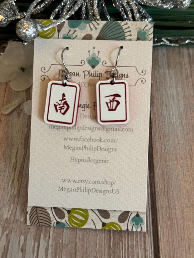 May include: White rectangular earrings with red Chinese characters, hanging from silver hooks. The earrings are displayed on a card with the text "Megan Philip Designs" and contact information.