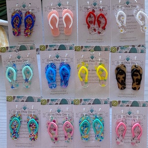 May include: Twelve pairs of colorful flip-flop earrings. Each pair is made of clear acrylic with a different patterned design. The earrings are displayed on white cards with the text "Megan Philip Designs" and a website address.