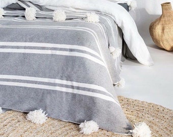 May include: A gray and white striped throw blanket with white pom poms. The blanket is on a bed with white bedding and pillows. There is a woven basket on the floor next to the bed.
