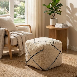 May include: A square, cream-colored ottoman with a shaggy texture and a black geometric design. The ottoman sits on a woven rug, near a wooden side table with a potted plant and a beige armchair with a blanket.