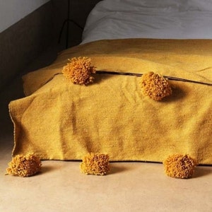 May include: A mustard yellow throw blanket with fluffy pom poms on a bed. The blanket is folded at the foot of the bed and there are pom poms on the floor.
