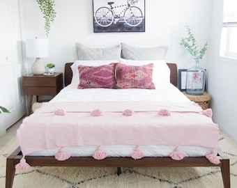 May include: A white bed with a pink throw blanket and a white and gray patterned rug. The bed has a wooden frame and a white headboard. There are two pillows on the bed, one with a pink and purple pattern and the other with a pink and red pattern. There is a black and white picture of a bicycle with flowers in the basket hanging on the wall above the bed.