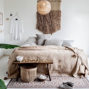 May include: A bedroom with a bed with a beige blanket and a wooden bench with a woven basket. The bed has a white headboard and a white throw pillow. There is a woven wall hanging above the bed and a light fixture hanging from the ceiling.