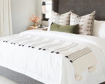 May include: A modern bedroom with a gray upholstered headboard and bed frame. The bed is covered with white linens, decorative pillows, and a cream-colored throw with black stripes and tassels. A small side table holds a vase of flowers.