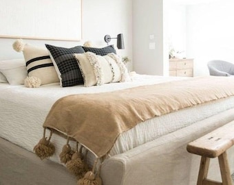 May include: A white bed with a tan throw blanket and a wooden stool. The bed has a white headboard and a white bedspread. The throw blanket has tassels on the edges. The stool is made of wood and has a natural finish.