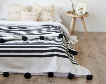 May include: A white and black striped throw blanket with black pom poms along the edges. The blanket is draped over a bed with white bedding and decorative pillows. A small wooden side table with a potted plant and a framed picture is in the background.